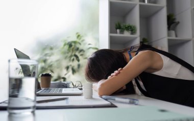 Genç bir ofis çalışanı yorgun görünüyor. Kafasını modern ofis ortamında masasına yaslıyor..