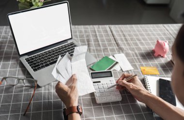 Finansal belgeleri ve faturaları incelerken, masada dizüstü bilgisayarı ve kumbarası olan, etkin bir bütçeyi vurgulayan odaklanmış bir adam..