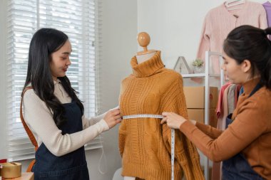 Two professional female designers meticulously measuring a cozy knitted sweater on a mannequin, working together in a trendy fashion studio. clipart