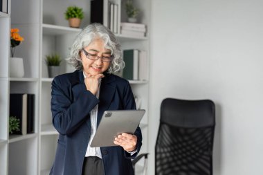 Gözlüklü kıdemli bir iş kadını tabletteki belgeleri neşeyle inceliyor, parlak bir ofis alanında düşünceli bir ifade sergiliyor..