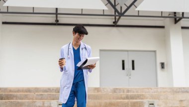 Genç bir doktor hastanenin dışında duruyor, hasta vakalarını inceliyor, modern sağlık hizmetlerini ve mesleki bağlılığı simgeliyor..