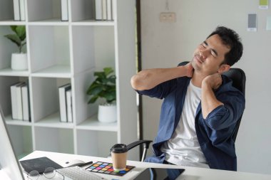Genç bir tasarımcı masa başında çalışırken boyun ağrısını hisseder, modern ofis ortamında ergonomik zorlukları vurgular.