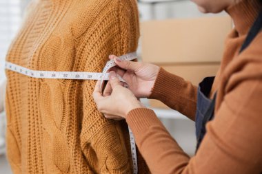 A dedicated woman measuring a cozy cable-knit sweater on a mannequin, ensuring the perfect fit in a stylish and organized fashion workshop. clipart