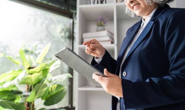 A senior businesswoman joyfully checking her documents on a tablet, standing beside lush indoor plants in a well-lit office space. clipart