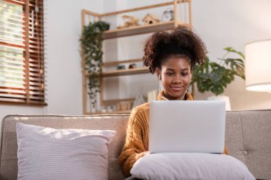 Genç Afrikalı bir kadın rahat rahat rahat evden çalışır, bir kanepede dizüstü bilgisayarla oturur, rahat ve modern bir oturma odası ortamıyla çevrilidir..
