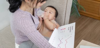 Kendini adamış bir anne bir iş planını incelerken bebeğini biberonla besler, rahat bir ev ortamında ebeveynlik ve mesleki sorumluluklarını dengeleme yeteneğini gösterir..