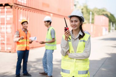 Yansıtıcı güvenlik yeleği giymiş kendine güvenen bir kadın inşaat yöneticisi telsiz kullanırken gülümsüyor, planlarını analiz ederken ekibini denetliyor ve proje detaylarını şantiyede tartışıyor..