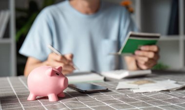 Finansal durumunu yönetmeye kendini adamış bir adam, elinde bütçe notları ve bir kalem, önünde pembe bir kumbara, birikimlerini ve finansal farkındalığını simgeliyor..