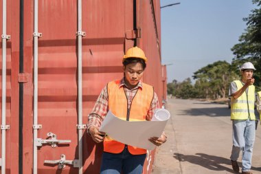 Turuncu güvenlik yeleği giyen genç bir inşaat mühendisi bir şantiyenin planlarını incelerken, sarı yelekli bir meslektaşı sözlü talimatlar veriyor..