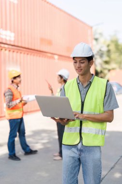 Sarı güvenlik yeleği giyen bir inşaat işçisi proje yönetimi için dizüstü bilgisayar kullanırken, takım arkadaşları da nakliye konteynırlarıyla çevrili inşaat alanının planlarıyla ilgili bir tartışmaya giriştiler..