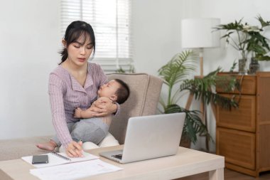 Kendini işine adamış bir anne, bebeğini yetiştirirken dizüstü bilgisayarıyla çalışıyor, ebeveynlik ve profesyonelliğin zorluklarını gösteriyor..