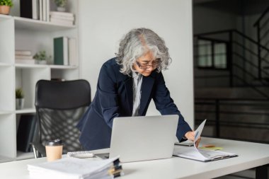 Deneyimli bir iş kadını, modern ofis ortamında organize evrak ve kahve ile çevrili bir dizüstü bilgisayara odaklanarak belgeleri etkili bir şekilde kontrol eder..