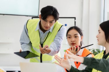 Güvenlik yeleği giymiş bir inşaat yöneticisi, bir tabletle ekiple temas kuruyor, bir işbirliği oturumu sırasında proje planları ve yönergeler hakkında ayrıntılı bir tartışma yürütüyor..