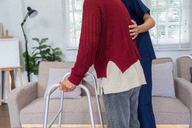 Şefkatli bir hemşire yaşlı bir adama rahat bir oturma odasında yürüteç kullanarak rehabilitasyon sırasında güvenliğini ve konforunu sağlamasına yardım eder..