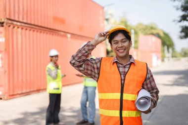 Canlı bir güvenlik yeleği ve sağlam bir şapkası olan mutlu bir inşaat işçisi, arka planda bir konteynır deposunda iş birliği yapan meslektaşlarıyla birlikte, güvenilir planlar tutuyor..