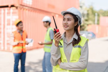 Sağlam bir şapka ve güvenlik yeleği giyen odaklanmış bir kadın inşaat denetçisi, arka planda canlı bir nakliye konteynırının arka bahçesinde ekip üyeleriyle çarpışırken bir telsiz tutuyor..