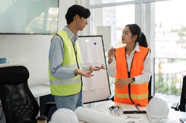 Proje detayları ve stratejileri üzerine odaklanmış bir tartışmada yer alan iki inşaat çalışanı, çağdaş ofis ortamında takım çalışması sergiliyor.