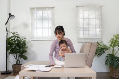 Odaklanmış bir anne, hem ailesine hem de işine bağlılığını gösteren bir dizüstü bilgisayarla bebeğini desteklerken ev ofisinde sorumluluklarını dengeler..