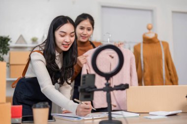 Two women joyfully recording details while showcasing a stylish pink sweater, carefully organizing their online clothing business in a vibrant workspace. clipart