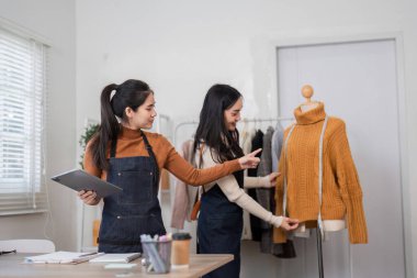 Parlak bir stüdyoda işbirliği yapan iki moda tasarımcısı, bir tanesi tablet tutarken giyilen rahat bir kazağın tasarım detaylarını analiz ediyor..