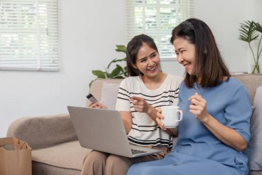 İki neşeli kadın internetten alışveriş yaparken keyifli bir anı paylaşıyor. Biri elinde bir kredi kartı, diğeri de bardaktan yudumluyor..