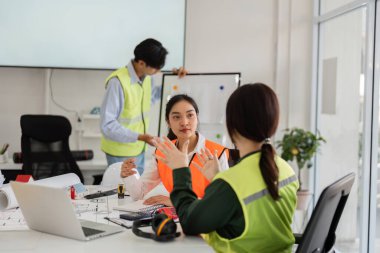 Bir mimar ve mühendis inşaat stratejileri hakkında dinamik bir konuşma yaparken, arka planda tasarımcı ve çağdaş bir ofiste planları gözden geçirir..
