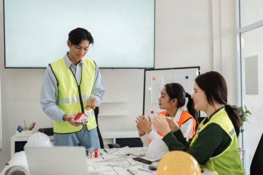 Çeşitli bir inşaat ekibi, bir meslektaşını coşkuyla alkışlıyor. Minyatür bir bina modeli sunuyor, canlı modern bir ofiste takım çalışması sergiliyor..