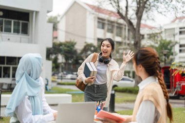 Neşeli, çok kültürlü bir öğrenci, kitap tutarken ve dışarıda, üniversite kampüsü ortamında kulaklık takarken arkadaşlarını selamlıyor..
