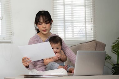 Bebeği modern bir ofis ortamında rahat bir şekilde kucağında otururken, odaklanmış bir anne belgeleri inceliyor..