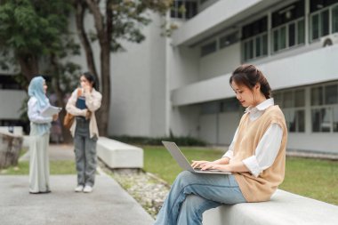 Çok kültürlü bir kadın dışarıda dizüstü bilgisayarla ders çalışmaya odaklanırken, arkadaşları üniversite ortamında sohbet ediyorlar..