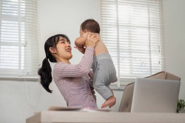 Neşeli bir anne, evinde oturmuş neşeli bağlarını kutlarken gülümseyerek bebeğini kaldırıyor..