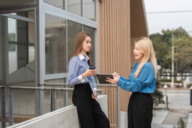 Çağdaş bir çalışma alanında bir tablet ve bir kahve bardağı tutarken, iki farklı iş kadını fikir alışverişinde bulunarak iş birliği yapıyor..