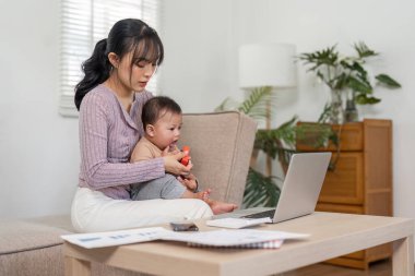 Bir anne, bebeğini kucağında tutarken ve rahat bir ev ortamında evrak işlerini yönetirken, modern ebeveynliği sergiliyor..