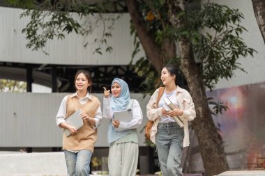 Üç üniversite öğrencisinden oluşan çeşitli bir grup, biri tesettür içinde, dizüstü bilgisayarlarını ve kitaplarını tartışırken ve taşırken kampüste yürüyorlar..