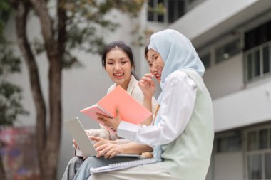 Açık havada ders çalışan, gülümseyen, notlarını paylaşan ve üniversite ortamında fikirleri tartışan iki farklı genç kadın..