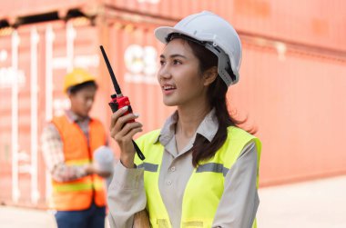 Sert bir şapka ve güvenlik yeleği giyen kendine güvenen bir kadın inşaat yöneticisi bir konteynır deposundaki açık hava projesi sırasında ekibiyle birlikte telsiz kullanıyor..