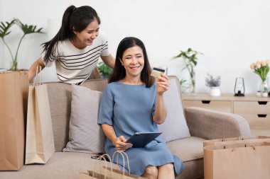 Mutlu kadınlar bir koltukta birlikte otururlar, biri kredi kartı taşır ve diğeri de tabletle online alışveriş deneyimlerinin tadını çıkarırken gizlice gülümser..