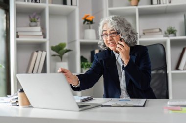 Yaşlı bir iş kadını bir telefon görüşmesi yapıyor ve aktif olarak laptopuyla çalışıyor. Etkili iletişim kuruyor ve şık ofis düzeninde birden fazla iş yapıyor..