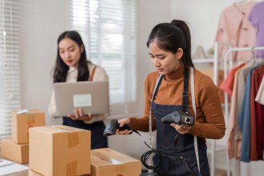Odaklanmış iki kadın, çevrimiçi satıcı, paketlenmiş siparişlerle dolu şık bir giyim mağazasında dizüstü bilgisayardaki verileri dikkatle incelerken barkod tarayıcılarıyla envanteri kontrol ediyorlar..