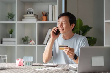 Masada oturan neşeli bir adam, elinde bir kredi kartı ve akıllı bir telefon, evde online alışveriş yaparken bir telefon görüşmesi yapıyor..