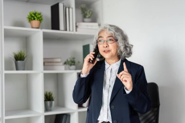 Önemli bir telefon görüşmesi yapan üst düzey bir işletmeci, kitap rafları ve yeşilliklerle süslenmiş modern bir ofiste güven içinde dururken kilit noktaları vurguluyor..