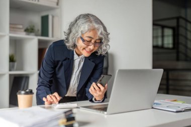 Yaşlı bir iş kadını, çağdaş bir ofis ortamında dokümanlar ve dizüstü bilgisayarla çevrili akıllı telefonunu kontrol ederken bir anlık pozitifliğin tadını çıkarıyor..