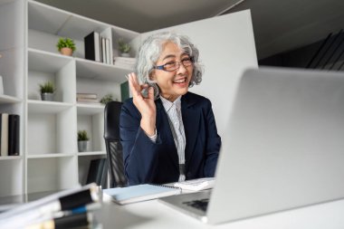 Neşeli, yaşlı bir iş kadını zarif ofisinde yaptığı bir video görüşmesi sırasında gülümsüyor ve onay veriyor, pozitifliğini ve nişanını gösteriyor..