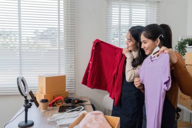 İki kadın, neşeli bir çalışma alanında online satışlarına hazırlanırken kırmızı bir kazak ve mor bir bluz sunarak neşe içinde kıyafet ürünlerini sergiliyorlar..