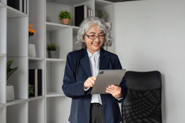 Gri saçlı üst düzey bir işletme yöneticisi tabletle mutlu bir şekilde iletişim kuruyor. Çağdaş bir ofis alanında güvenle duruyor. Bitkilerle dolu ve organize raflarla dolu..