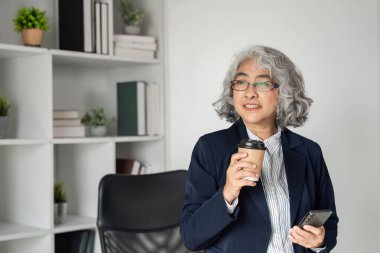 Kendine güvenen üst düzey bir işletme yöneticisi, şık bir ofis yerinde akıllı bir telefon tutarken kahvesinin tadını çıkarıyor, profesyonel ve cana yakın tavrını sergiliyor..