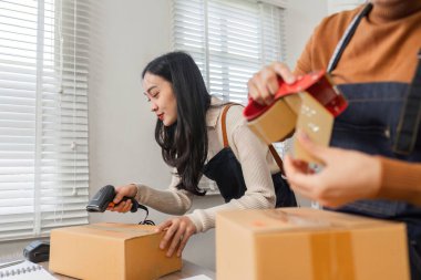 A diverse couple scanning packages with a barcode scanner while preparing online orders for shipment in their organized boutique workspace. clipart