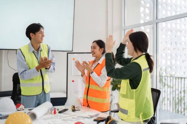 Bir mimar, mühendis ve tasarımcıdan oluşan neşeli bir inşaat ekibi, çağdaş ofis ortamında bir proje kilometre taşını başarma şerefine el çırpıyor..