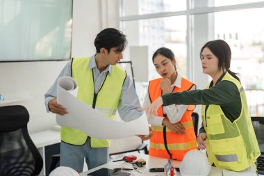 Çeşitli bir inşaat ekibi proje planlarını işbirliği içinde gözden geçirir ve parlak, modern bir ofiste planları tartışır, takım çalışmasını gösterir ve odaklanır..