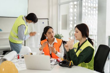 Bir mimar, mühendis ve tasarımcı ile birlikte bir mimari ekibi, şık bir ofis ortamında iş birliği yaparken inşaat planları hakkında ayrıntılı bir tartışmaya girişiyor..